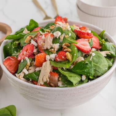 A bowl of spinach salad with sliced strawberries, shredded chicken, crumbled cheese, and pecans on a marble surface, surrounded by small bowls and utensils.
