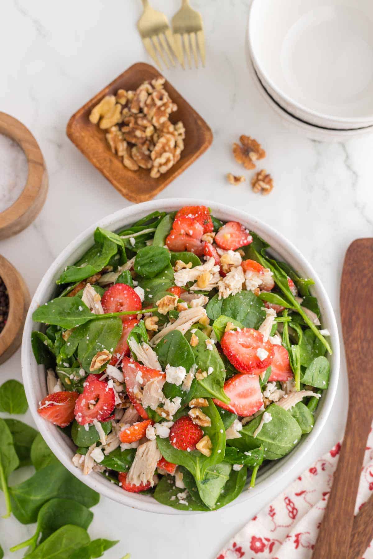 A bowl of spinach salad with strawberries, shredded chicken, crumbled cheese, and walnuts on a marble surface, with utensils and salad ingredients nearby.