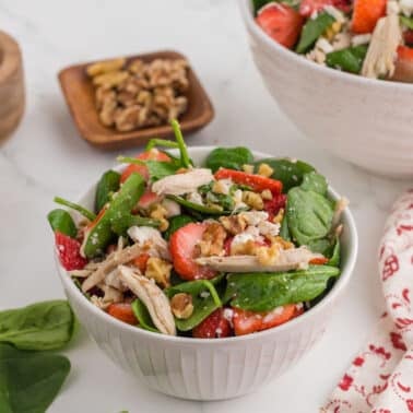 A bowl of salad with spinach, shredded chicken, strawberries, walnuts, and feta cheese sits on a white surface next to spices and a patterned napkin.