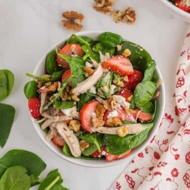 A bowl of spinach salad with shredded chicken, sliced strawberries, walnuts, and feta cheese, placed on a white surface next to fresh spinach leaves and a patterned cloth.