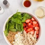 A bowl containing fresh spinach, sliced strawberries, shredded chicken, crumbled feta cheese, and chopped walnuts. Olive oil, vinaigrette, salt, and pepper are arranged nearby.