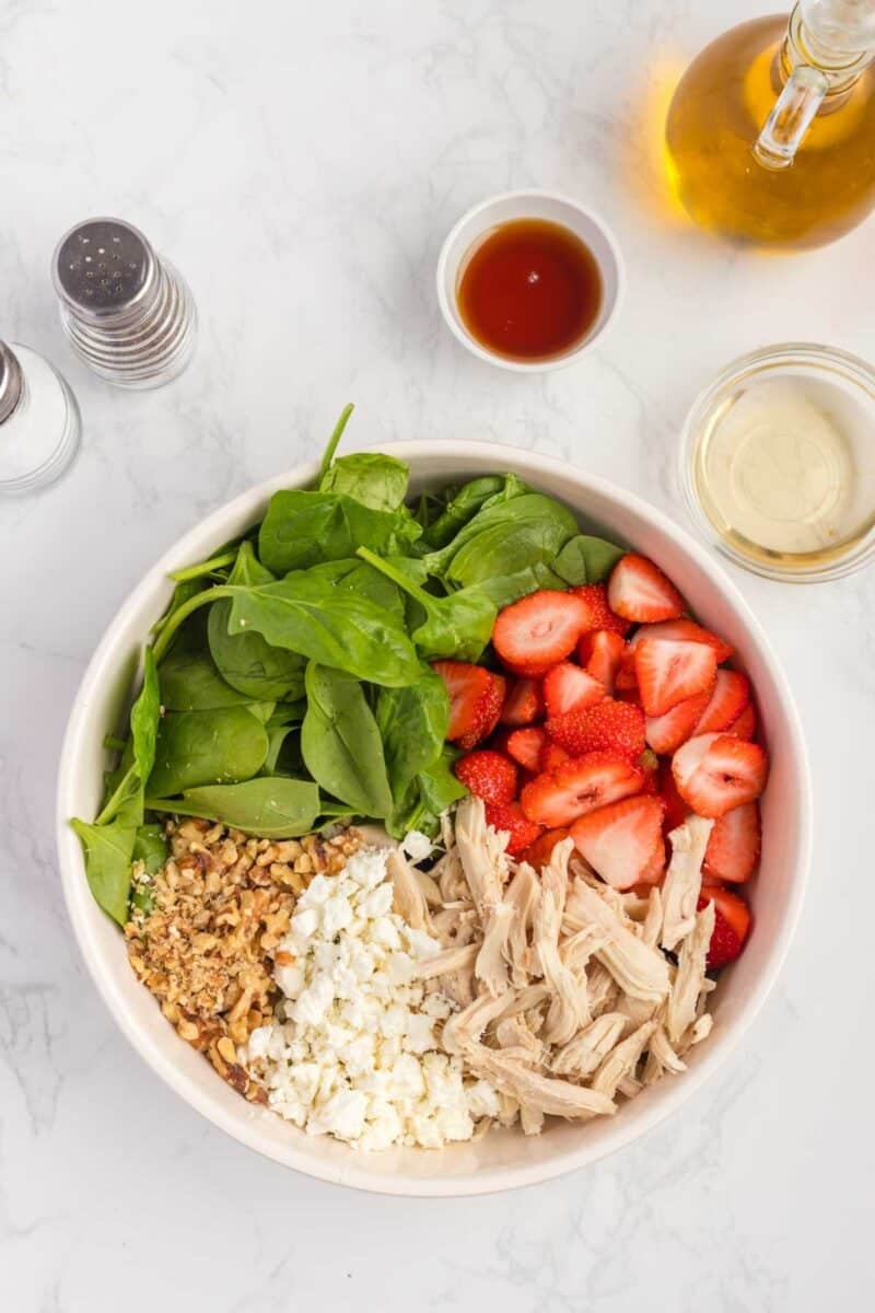 A bowl containing fresh spinach, sliced strawberries, shredded chicken, crumbled feta cheese, and chopped walnuts. Olive oil, vinaigrette, salt, and pepper are arranged nearby.