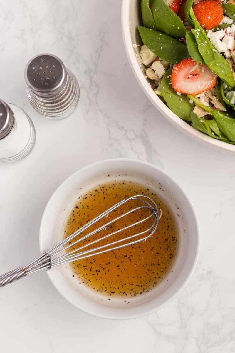 A bowl of vinaigrette with a whisk sits next to a spinach and strawberry salad; salt and pepper shakers are nearby on a marble countertop.