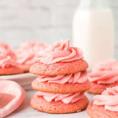 Three pink frosted strawberry sugar cookies are stacked in the foreground, with more cookies, a pink cloth, and a bottle of milk in the background.
