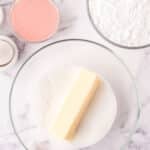 A glass bowl with a stick of butter and granulated sugar, surrounded by bowls of pink sugar, powdered sugar, and eggs on a marble surface.