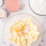 A mixing bowl with butter, sugar, and an egg, surrounded by bowls of powdered sugar, pink powder, and eggs on a marble surface.