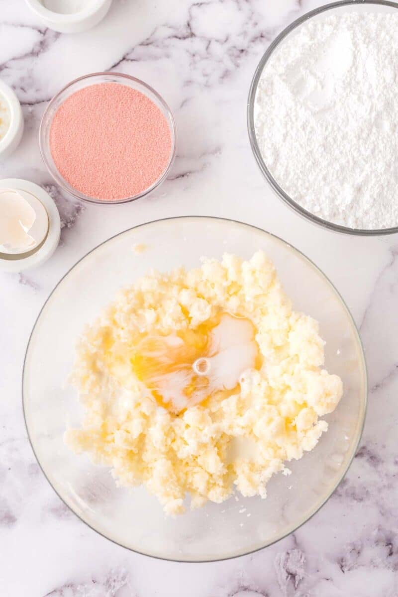 A mixing bowl with butter, sugar, and an egg, surrounded by bowls of powdered sugar, pink powder, and eggs on a marble surface.