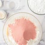 A glass bowl with white and pink powdered ingredients sits on a marble surface, surrounded by bowls of powdered sugar and two egg shells.