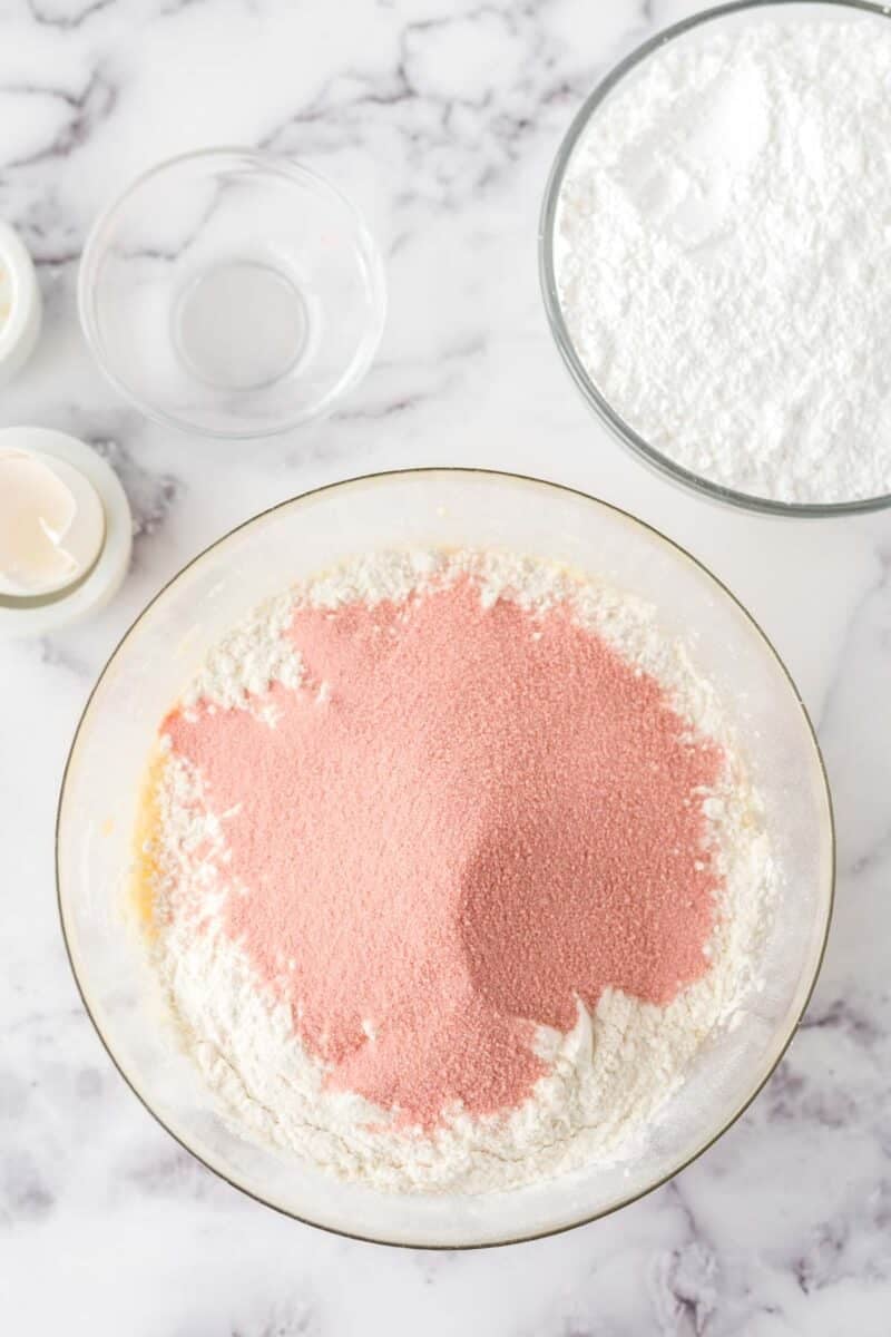 A glass bowl with white and pink powdered ingredients sits on a marble surface, surrounded by bowls of powdered sugar and two egg shells.