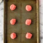 A metal baking tray with six evenly spaced scoops of pink cookie dough, ready to be baked. The tray is on a white marble surface.