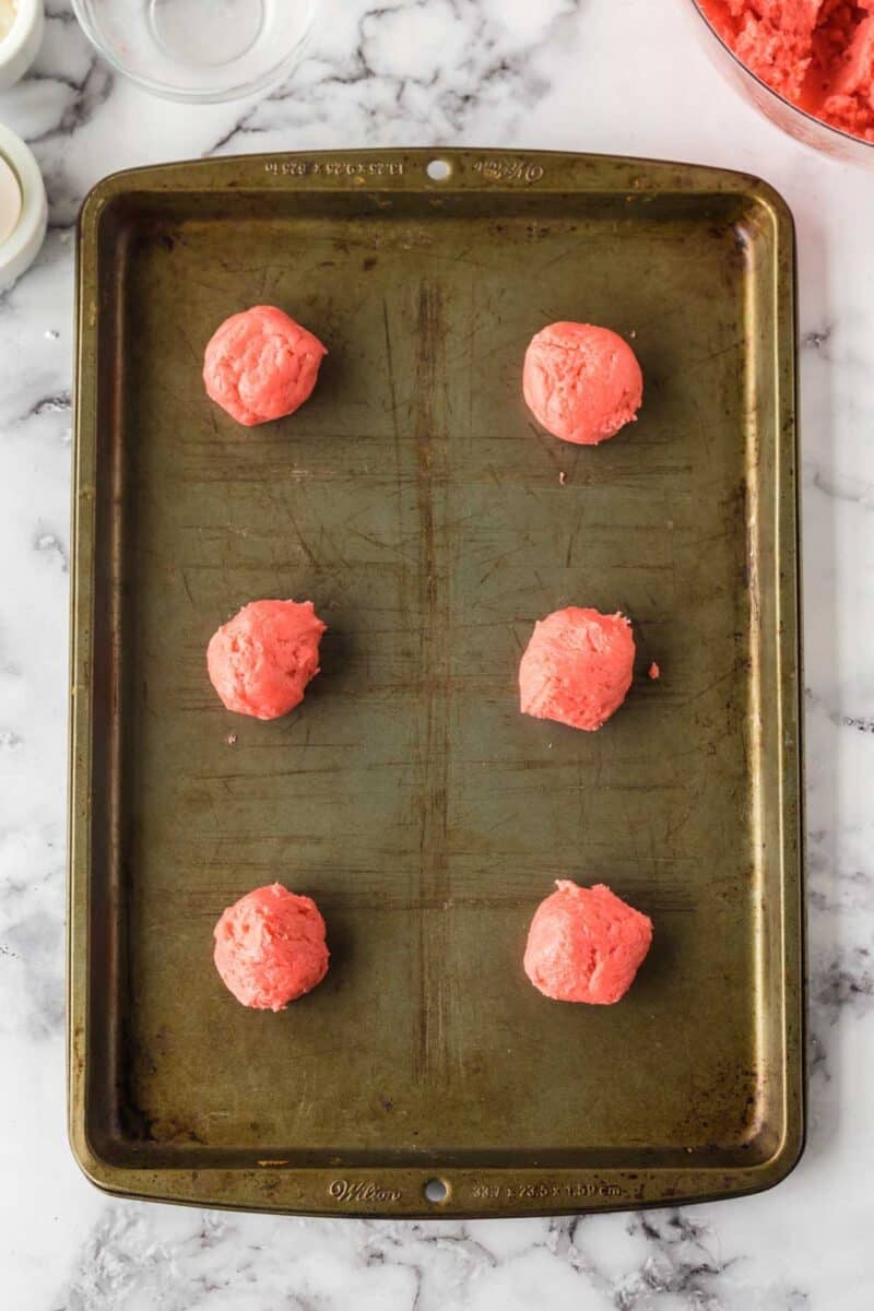 A metal baking tray with six evenly spaced scoops of pink cookie dough, ready to be baked. The tray is on a white marble surface.