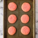 Six round, pink cookies are evenly spaced on a metal baking sheet placed on a marble countertop.