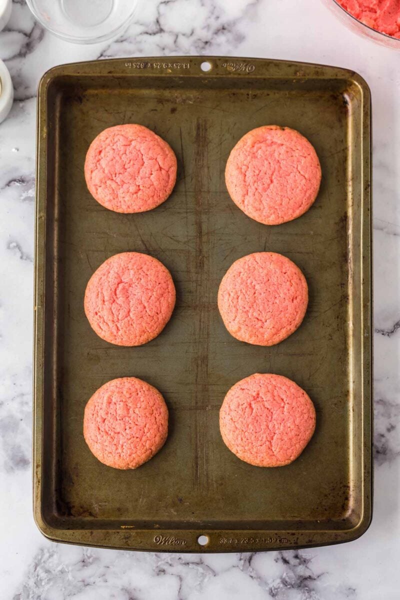 Six round, pink cookies are evenly spaced on a metal baking sheet placed on a marble countertop.