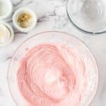 A glass bowl filled with pink frosting sits on a marble surface, surrounded by empty glass bowls and small containers of ingredients.