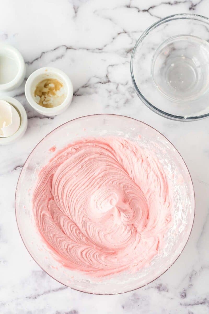 A glass bowl filled with pink frosting sits on a marble surface, surrounded by empty glass bowls and small containers of ingredients.