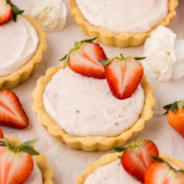 Mini tart shells filled with creamy pink filling, each topped with halved strawberries, are arranged on a light surface with whole strawberries and white flowers nearby.