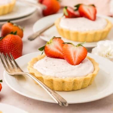 A small tart with pink filling, topped with sliced strawberries, is served on a white plate with a fork. Whole and sliced strawberries are in the background.