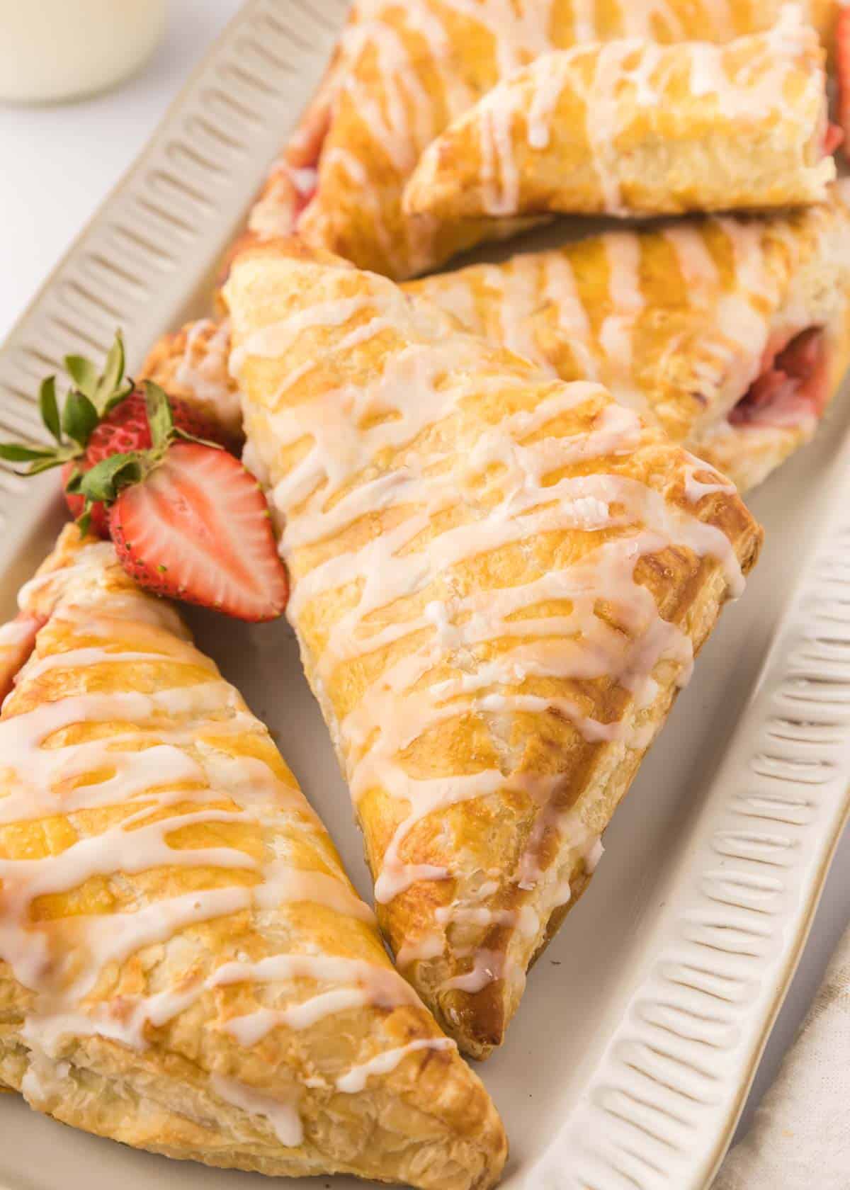 Triangular strawberry turnovers with icing drizzle are arranged on a rectangular white plate, garnished with a halved strawberry.