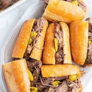 Several sandwiches filled with shredded chicago italian beef and pepperoncini peppers are arranged on a white platter.