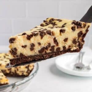A close-up of a slice of chocolate chip cheesecake with a chocolate crust being served from a glass pie dish.