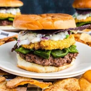 A hamburger topped with green peppers, a breaded fried patty, creamy sauce, and a blue corn chip on a white plate, with potato chips on the side.