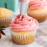 A piping bag is frosting a vanilla cupcake with pink strawberry buttercream icing; other cupcakes and a strawberry are visible in the background.