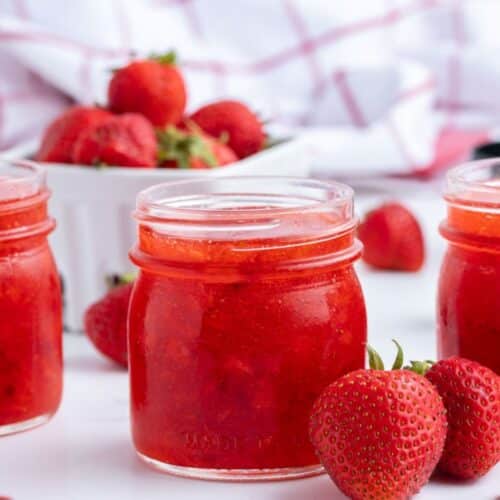 Three jars of strawberry jam sit on a white surface, with fresh strawberries and a container of strawberries in the background.