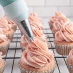 A piping bag applies strawberry frosting with jam onto a cupcake, with several frosted cupcakes on a wire rack in the background.