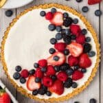 A berry tart with a cream filling is topped with a crescent of fresh strawberries, raspberries, blackberries, and blueberries, sitting on a wooden surface.