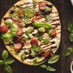 Flatbread pizza topped with cherry tomatoes, mozzarella, fresh basil leaves, pesto, and a drizzle of balsamic glaze on a dark wooden surface.