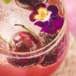 A glass of pink sparkling drink with ice, fresh cherries, and a purple edible flower garnish.