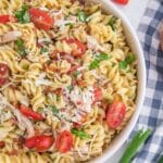 A bowl of rotini pasta salad with cherry tomatoes, shredded chicken, chopped spinach, bacon, and grated cheese, next to a blue and white checkered napkin.