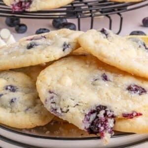 A plate of blueberry cookies with sugar crystals on top, white chocolate chips, and a cooling rack with more cookies in the background.