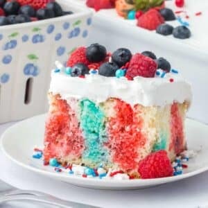 A slice of patriotic poke cake with red, white, and blue colors, topped with whipped cream, blueberries, raspberries, and sprinkles, on a white plate.