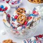 A glass bowl filled with white chocolate bark topped with pretzels, red and blue candies, and mini marshmallows. Other candy and snacks are visible in the background.