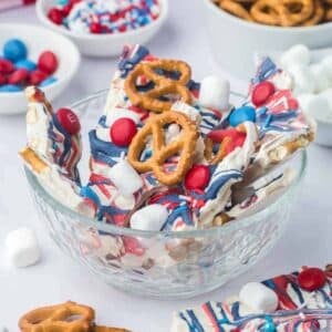 A glass bowl filled with white chocolate bark topped with pretzels, red and blue candies, and mini marshmallows. Other candy and snacks are visible in the background.