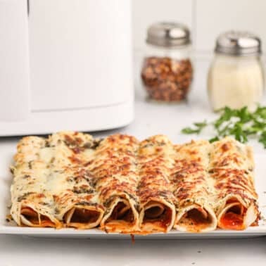 A plate of baked cheese-covered pizza rollups sits on a countertop, with a bowl of red sauce, spice jars, fresh herbs, and a bottle of olive oil in the background.