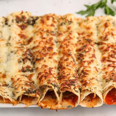 A plate of baked pizza rollups covered with melted cheese and herbs, with tomato sauce visible inside the tortillas.