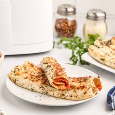 Two air fryer pizza roll-ups on a white plate, sprinkled with herbs, with a bowl of marinara sauce and ingredients like olive oil, spices, and parsley in the background.