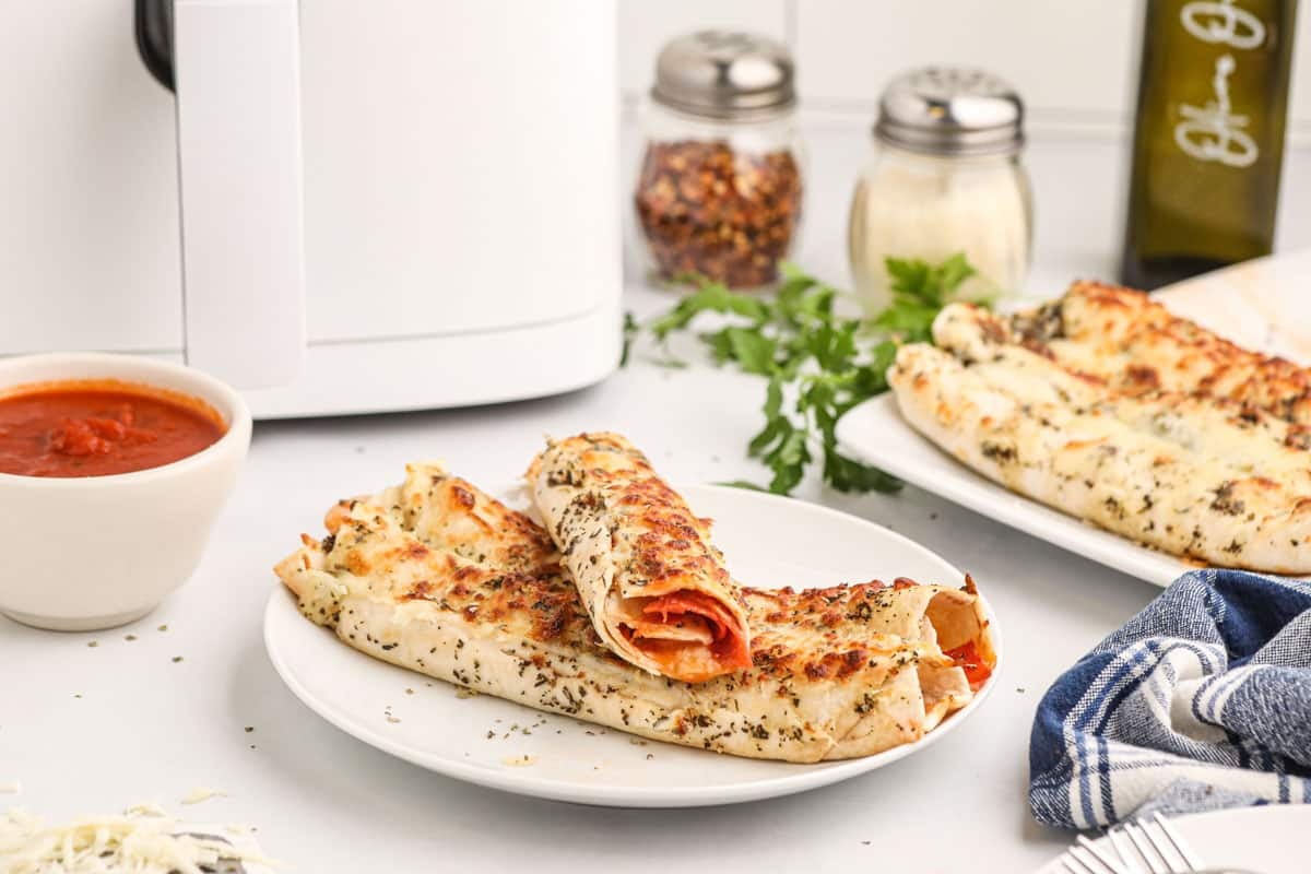 Two air fryer pizza roll-ups on a white plate, sprinkled with herbs, with a bowl of marinara sauce and ingredients like olive oil, spices, and parsley in the background.