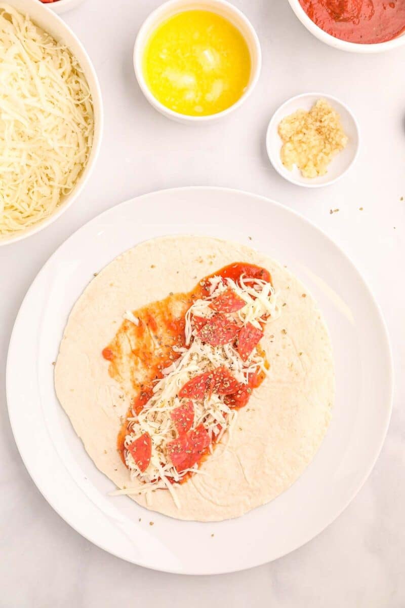 A tortilla on a white plate topped with tomato sauce, shredded cheese, pepperoni slices, and herbs, surrounded by small bowls of melted butter, garlic, cheese, and sauce.