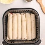 Six rolled tortillas are lined up in an air fryer basket. A bowl of shredded cheese, a bowl of seasoning, and a basting brush are placed nearby on a white surface.