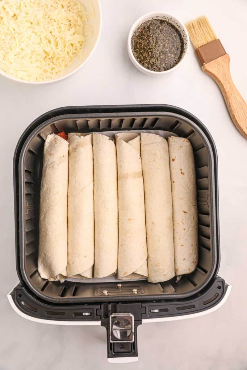 Six rolled tortillas are lined up in an air fryer basket. A bowl of shredded cheese, a bowl of seasoning, and a basting brush are placed nearby on a white surface.