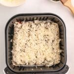 Air fryer basket with unbaked cheese-covered air fryer pizza rollups, a bowl of shredded cheese, and a small bowl of herb mixture with a brush on a white surface.