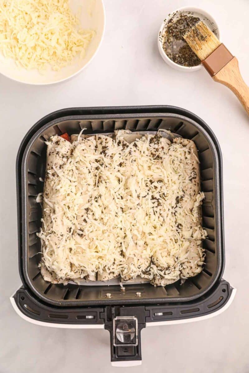 Air fryer basket with unbaked cheese-covered air fryer pizza rollups, a bowl of shredded cheese, and a small bowl of herb mixture with a brush on a white surface.
