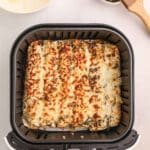 Cheesy air fryer pizza rollups topped with herbs are shown cooked in an air fryer basket, with a small bowl of shredded cheese and a basting brush with seasoning nearby.