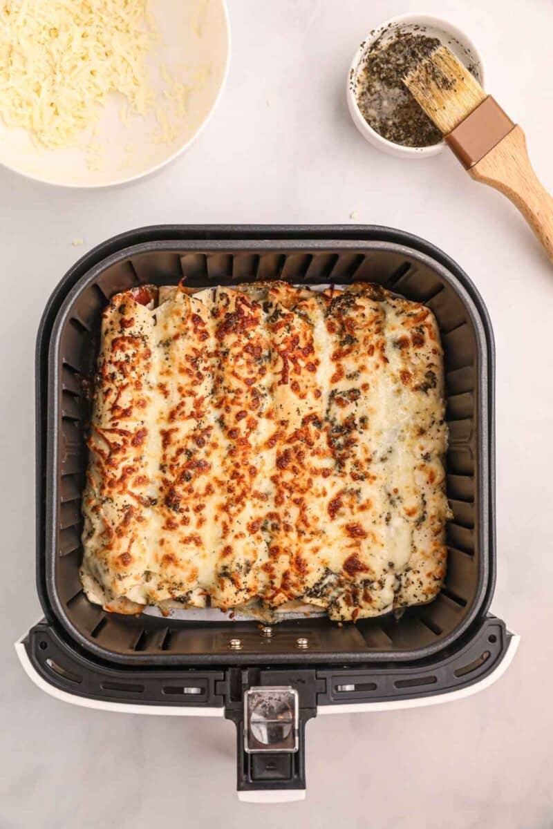 Cheesy air fryer pizza rollups topped with herbs are shown cooked in an air fryer basket, with a small bowl of shredded cheese and a basting brush with seasoning nearby.