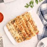 Air fryer pizza rollups sits on a white plate, topped with melted cheese and herbs, surrounded by a sauce bowl, cheese grater, utensils, and fresh parsley on a white surface.