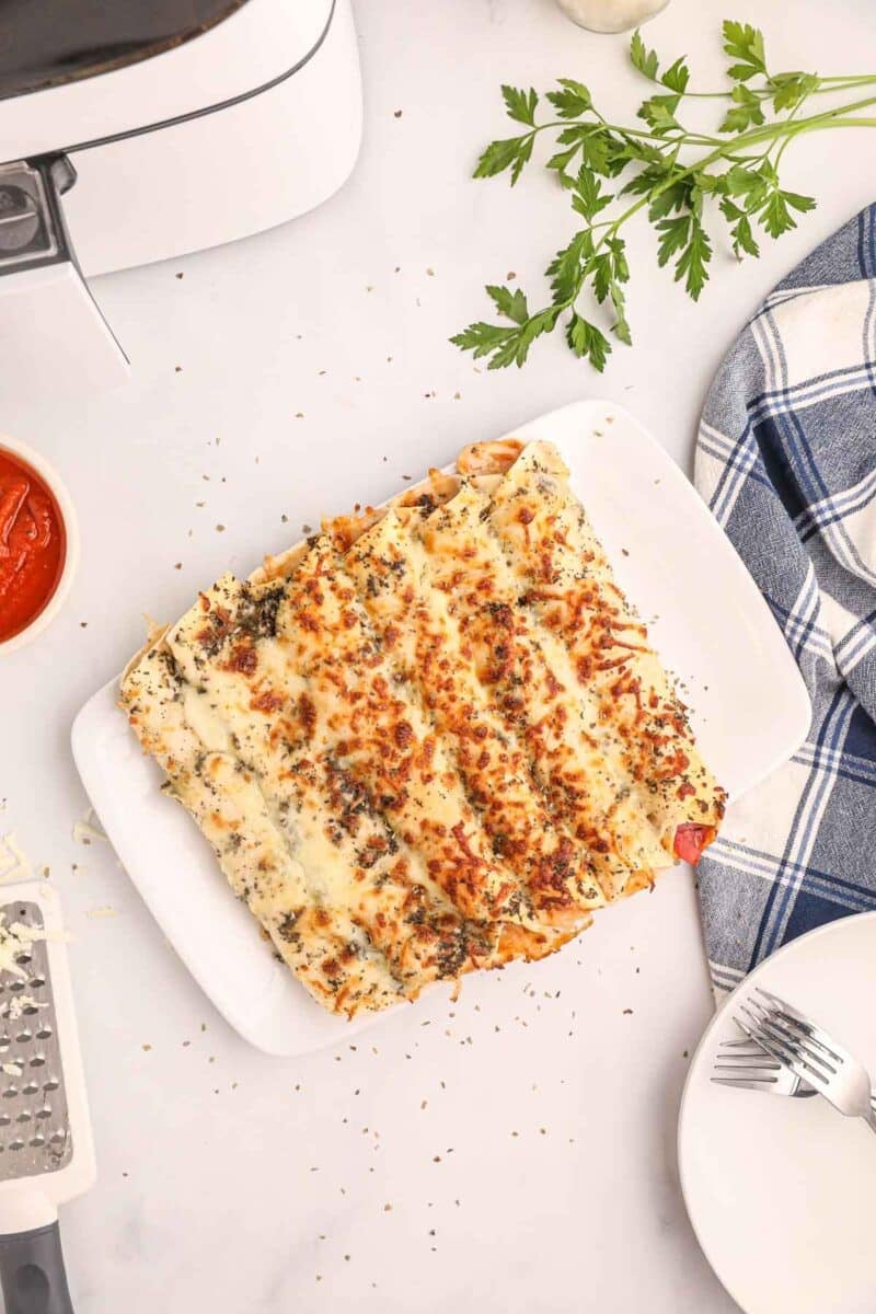 Air fryer pizza rollups sits on a white plate, topped with melted cheese and herbs, surrounded by a sauce bowl, cheese grater, utensils, and fresh parsley on a white surface.