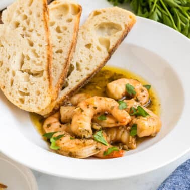 A white bowl contains New Orleans barbecue shrimp in a garlic-herb sauce, garnished with parsley, and served with two slices of rustic bread. Fresh parsley and more bread are nearby.